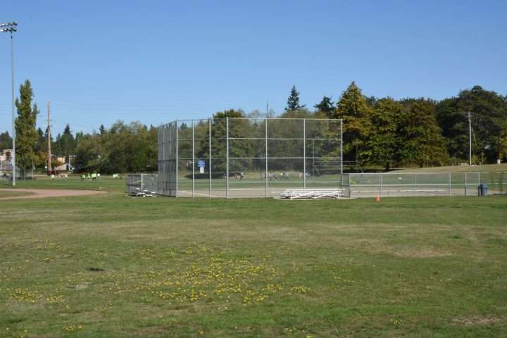Lakota Park. A wide view of an outdoor baseball field surrounded by tall chain-link fencing on a sunny day. Bleachers sit along the side of the field, and a few people wearing bright green shirts are gathered in the distance. The field is bordered by trees with early fall foliage, and the sky above is clear and blue.