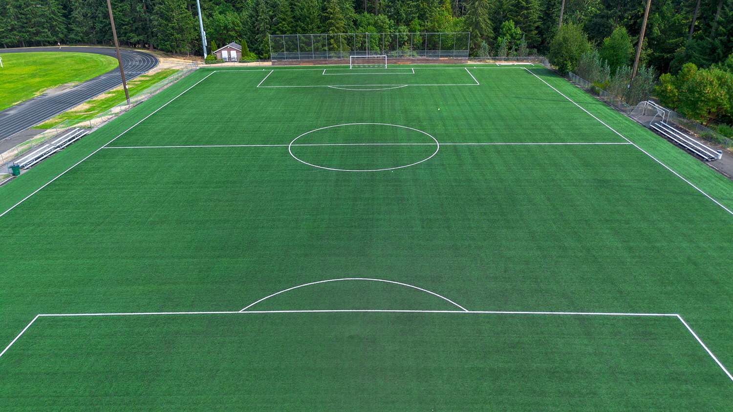 Elevated view of an empty full-size artificial turf soccer field with bright white field markings, goals at both ends, stadium lighting, aluminum bleachers along the sidelines, and a running track visible to the left. Dense evergreen trees border the facility in the background.