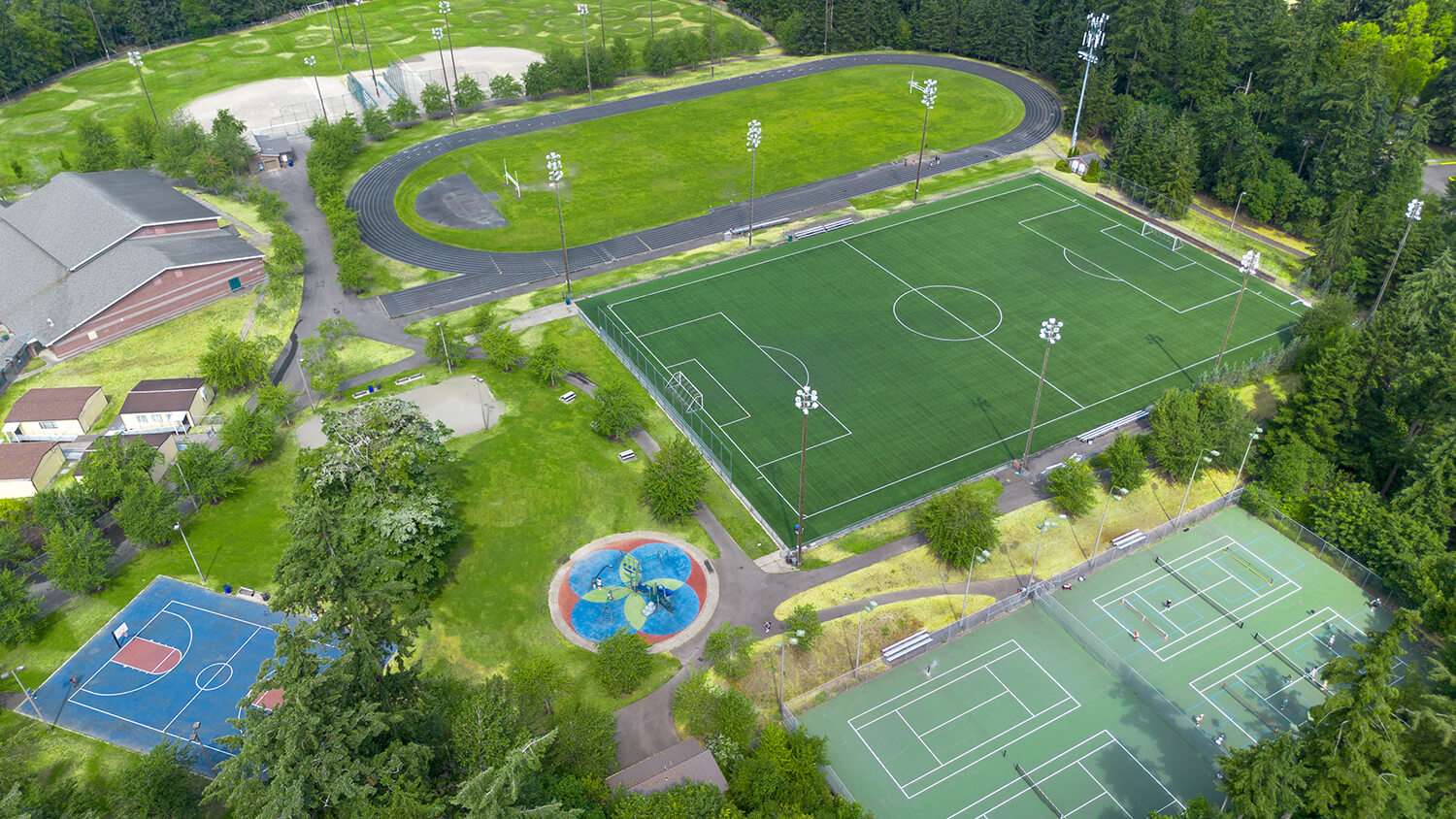 Aerial view of a Saghalie Park surrounded by dense trees. The facility features a full-size artificial turf soccer field with stadium lighting, an oval running track enclosing a grass infield, multiple tennis and pickleball courts, an outdoor basketball court, a colorful children's playground, walking paths, benches, and a large recreation building. Additional baseball diamonds are visible beyond the track.